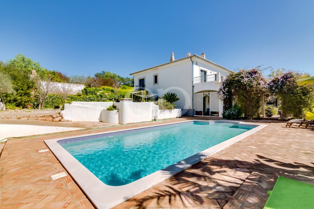 Moradia branca de dois andares com varanda com vista para uma piscina retangular de águas turquesa, um deck em terracota e um jardim exuberante sob um céu azul límpido, num pátio ensolarado.