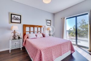 Quarto com uma cabeceira de madeira, uma colcha geométrica cor-de-rosa e duas mesas de cabeceira com tampo de vidro e candeeiros.