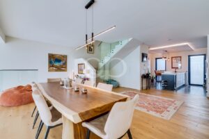 Un espace salon-salle à manger ouvert, avec une table en bois massif et des chaises beiges, un escalier à rampe en verre et des suspensions lumineuses modernes aux lignes épurées.