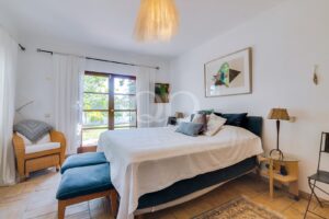 Chambre lumineuse avec un grand lit recouvert d'un couvre-lit blanc et d'une parure de lit bleu sarcelle, donnant sur des portes-fenêtres ouvrant sur le jardin.