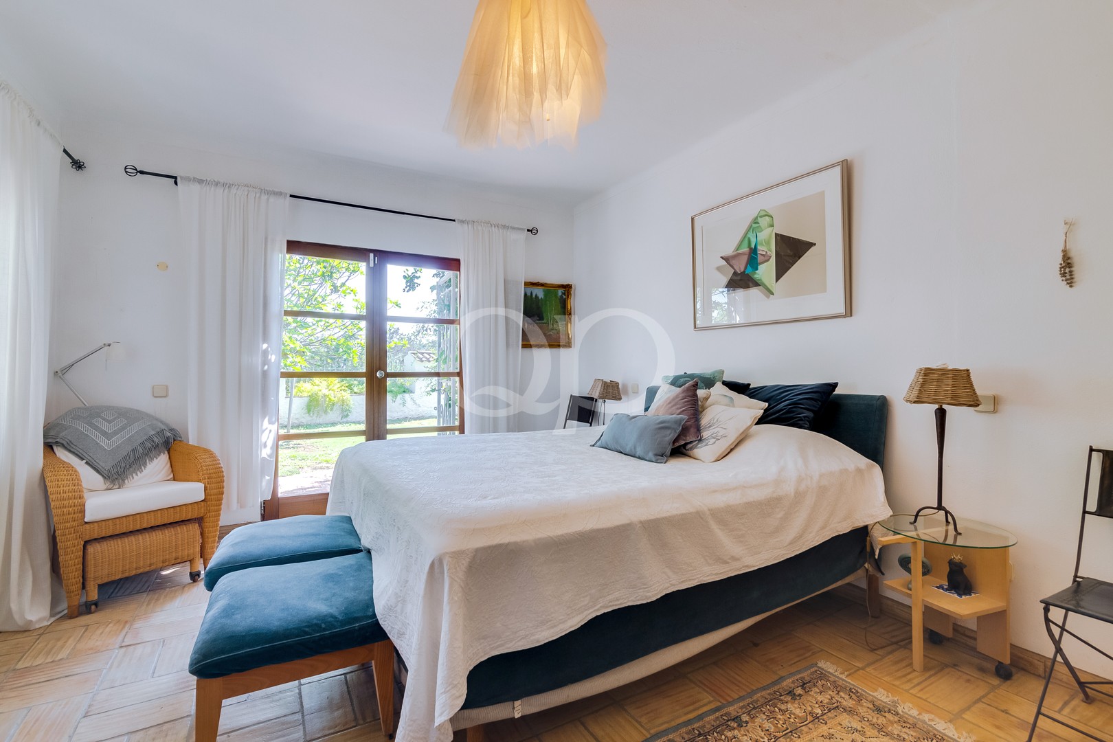 Chambre lumineuse avec un grand lit recouvert d'un couvre-lit blanc et d'une parure de lit bleu sarcelle, donnant sur des portes-fenêtres ouvrant sur le jardin.