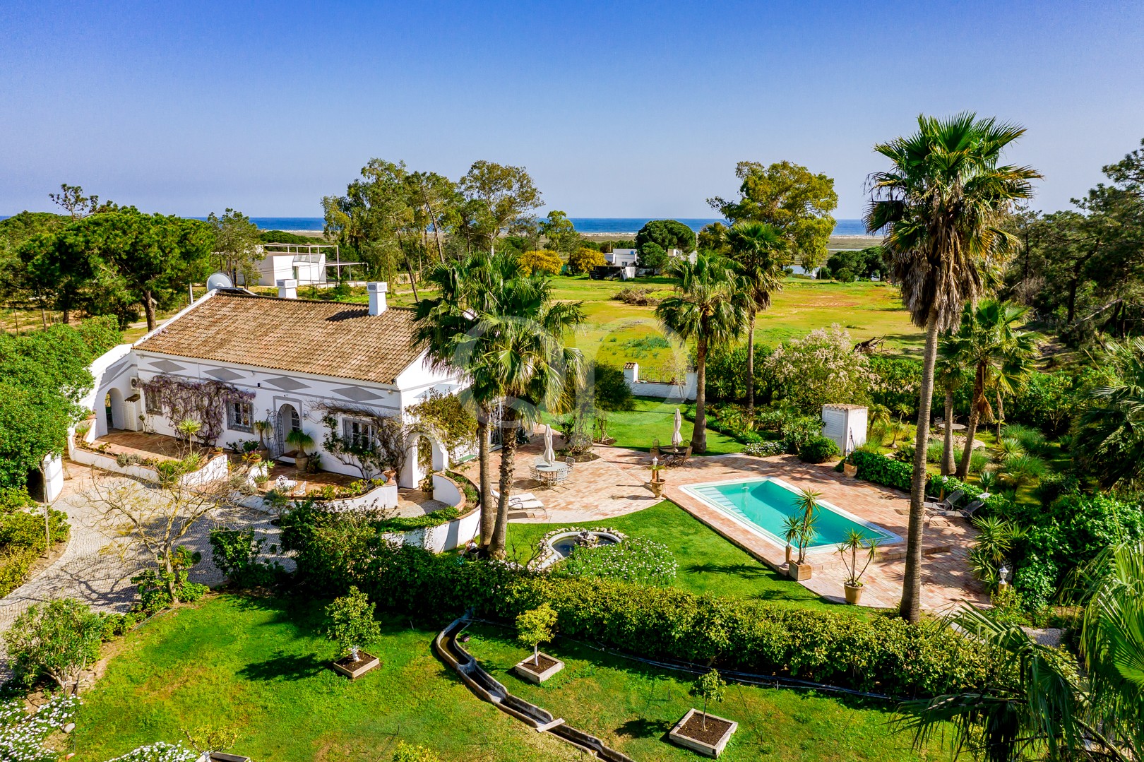 Villa blanche au toit en terre cuite, entourée de palmiers et dotée d'une piscine rectangulaire, nichée dans un jardin luxuriant avec vue sur la mer au loin.