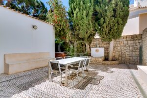 Sunny Mediterranean-style courtyard with a white wall, built-in bench, and a white dining table surrounded by six chairs on a cobblestone patio. Tall trees and a stone wall frame the background.