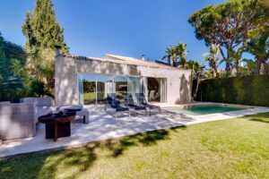 Modern single-story home with sliding glass doors, outdoor lounge chairs, and a rectangular pool in a sunny backyard.