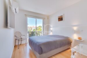 Bright modern bedroom with a large gray bed, white walls, and a balcony door showing palm trees outside amid sunny light.