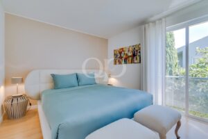 Bright bedroom with a light blue duvet, white upholstered bed, and abstract colorful wall art near a glass door.
