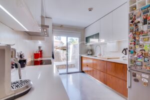 Bright modern kitchen with white cabinets, wooden lower drawers, and a glass sliding door to an outdoor patio; fridge covered in magnets on the right.