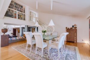 Bright dining room with a glass-topped table and six cream chairs on a floral rug, in a spacious open living area.
