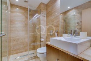 Modern beige marble bathroom with glass shower enclosure, white rectangular sink, wooden vanity, and toilet.