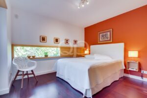 Bright modern bedroom with orange accent wall, white bed and headboard, bedside lamps, and a long window shelf with plants and bowls along the ledge on the left.