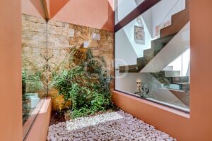 Indoor courtyard with stone wall, climbing greenery, white pebbles, and a glass-walled staircase area. They present a modern, warm-toned atrium interior.