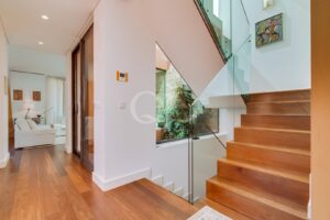 Modern entry hall with warm wood floor and a glass-railed wooden staircase; living room with white seating visible in the background.