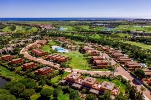 Aerial view of a coastal resort with tan, cube-shaped villas surrounding a central pool and expansive green lawns