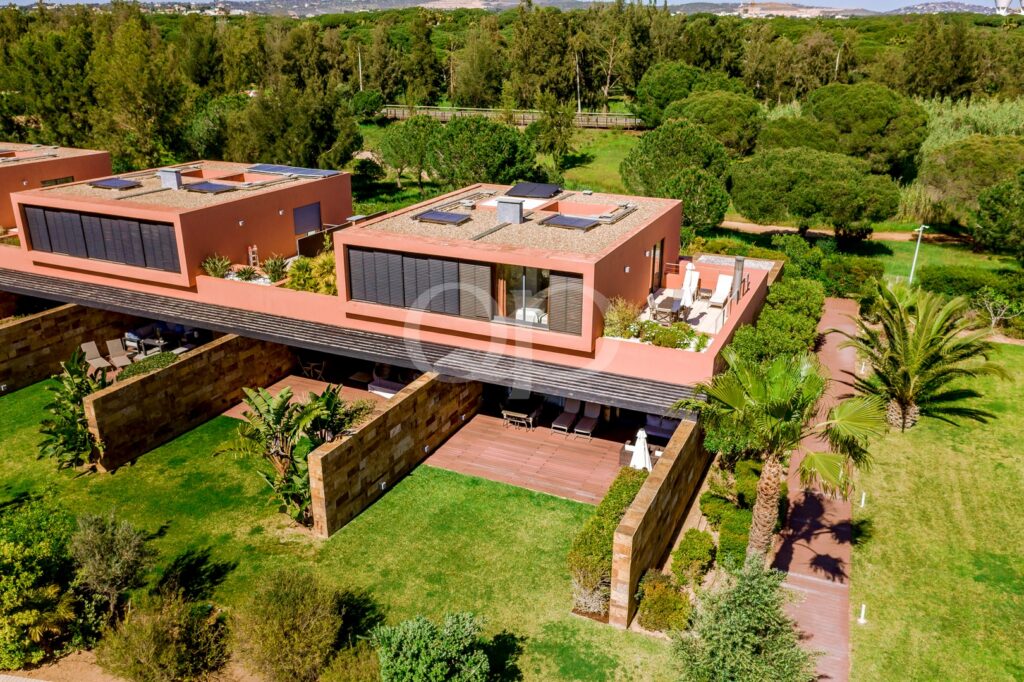 Aerial view of a terracotta villa with flat roofs, solar panels, large glass doors, and lush lawns surrounded by palm trees and patios.