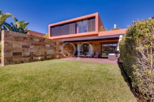 Backyard of a modern orange-framed house with a stone privacy wall, glass doors, and a tiled patio with dining furniture and lounge chairs in front of a green lawn.