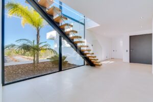 Interior minimalista e luminoso, com uma escadaria flutuante em madeira e um corrimão em vidro junto a uma parede de vidro do chão ao teto, com vista para as palmeiras no exterior.