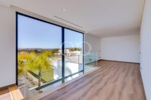 Sala de estar luminosa e moderna, com uma grande parede de vidro do chão ao teto e um corrimão de vidro com vista para um jardim ajardinado com palmeiras.