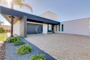 Casa moderna de dois andares com portas de garagem cinzentas escuras, paredes brancas, um pilar de pedra e uma palmeira numa entrada de automóveis de calçada de pedra I