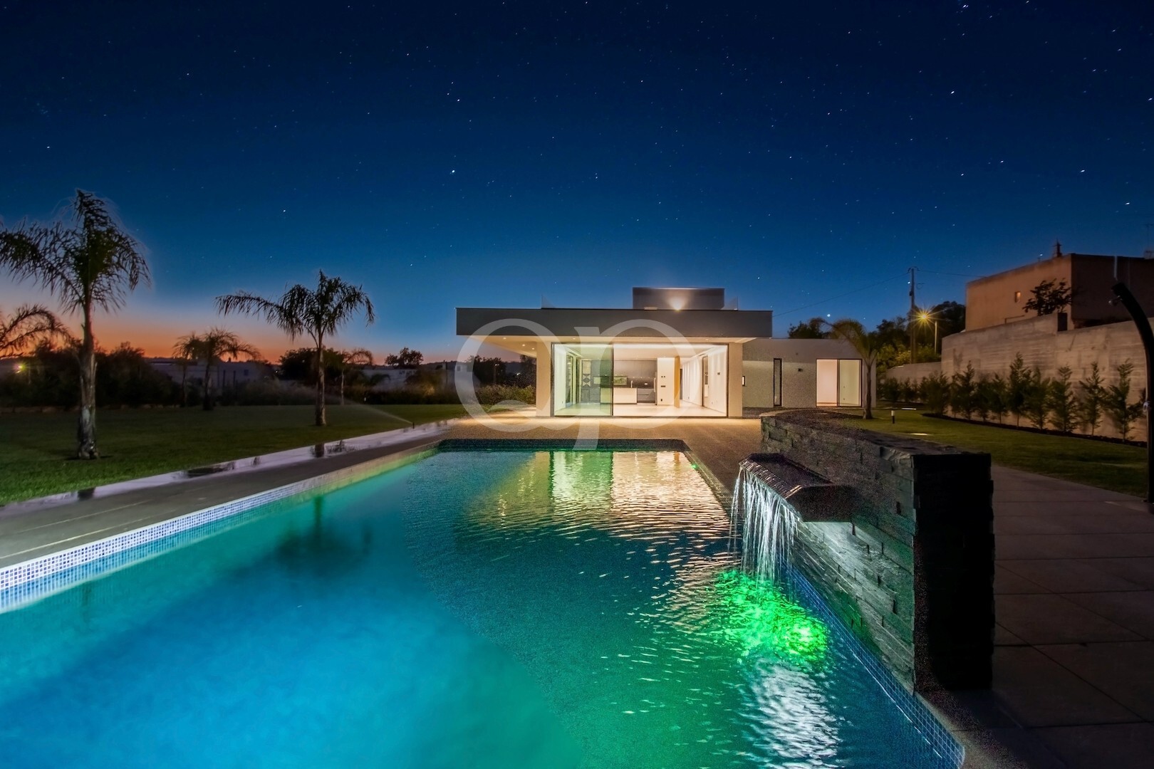 Casa moderna de um só piso com paredes de vidro, piscina iluminada à noite e palmeiras sob um céu estrelado.