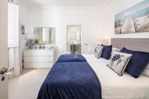 Bright coastal bedroom with white walls, navy bedding, and sea-themed pillows; doorway to a tiled bathroom.