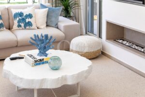 Cozy living room with a white round coffee table, blue coral sculpture, magazines, and a beige sofa with blue palm-patterned cushions.