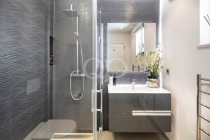 Modern grey bathroom with a glass shower enclosure, wavy textured tiles, and a dark grey vanity with a white sink and mirror.