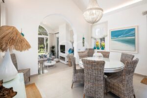 Bright coastal dining area with a glass round table and eight woven rattan chairs, a crystal chandelier, and a beach-themed painting on the wall; arched opening leads to a living room with a fireplace and large windows showing greenery.