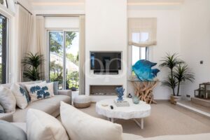 Bright living room with a beige sofa, blue palm-tree cushions, and a wall-mounted TV above a fireplace.