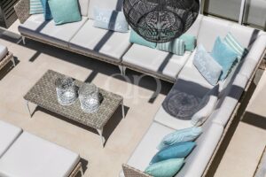 Top-down view of white cushioned outdoor sectional with teal and blue pillows around a glass-tiber coffee table and woven lanterns casting shadows above.