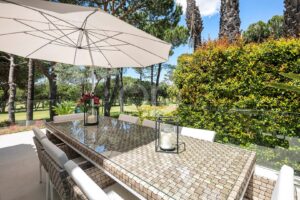 Outdoor patio dining area with a woven glass-top table, white chairs, and a large beige umbrella, surrounded by green hedges and trees outside a glass railing.