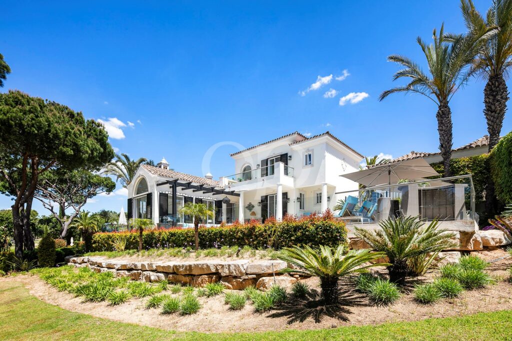 White Mediterranean-style villa with a poolside patio, palm trees, and a manicured garden under a bright blue sky