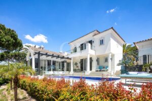 White Mediterranean-style villa with a glass-paneled pool, outdoor seating, and palm trees on a sunny day.