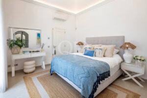 Bright, coastal-style bedroom with a large upholstered headboard, blue throw blanket, and palm-themed pillows on a white bed. Two white nightstands with wicker lamps flank the bed; a woven rug covers the floor.