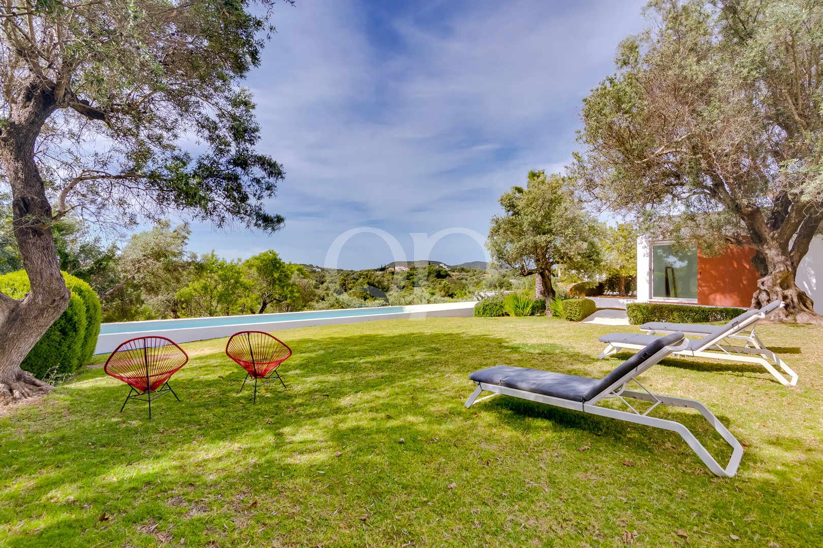 Une pelouse bordant une piscine rectangulaire, deux chaises en fil de fer rouges, des chaises longues blanches, des oliviers et un bâtiment blanc rehaussé de touches orange.