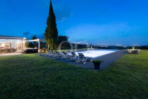 Dusk poolside with glass-walled house, row of lounge chairs, and a tall evergreen tree against a blue sky; distant city lights on the horizon.