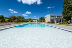 Ein weitläufiger Pool im Resort-Stil mit kristallklarem Wasser, weißer Terrasse, Liegestühlen und einer modernen Villa unter blauem Himmel.