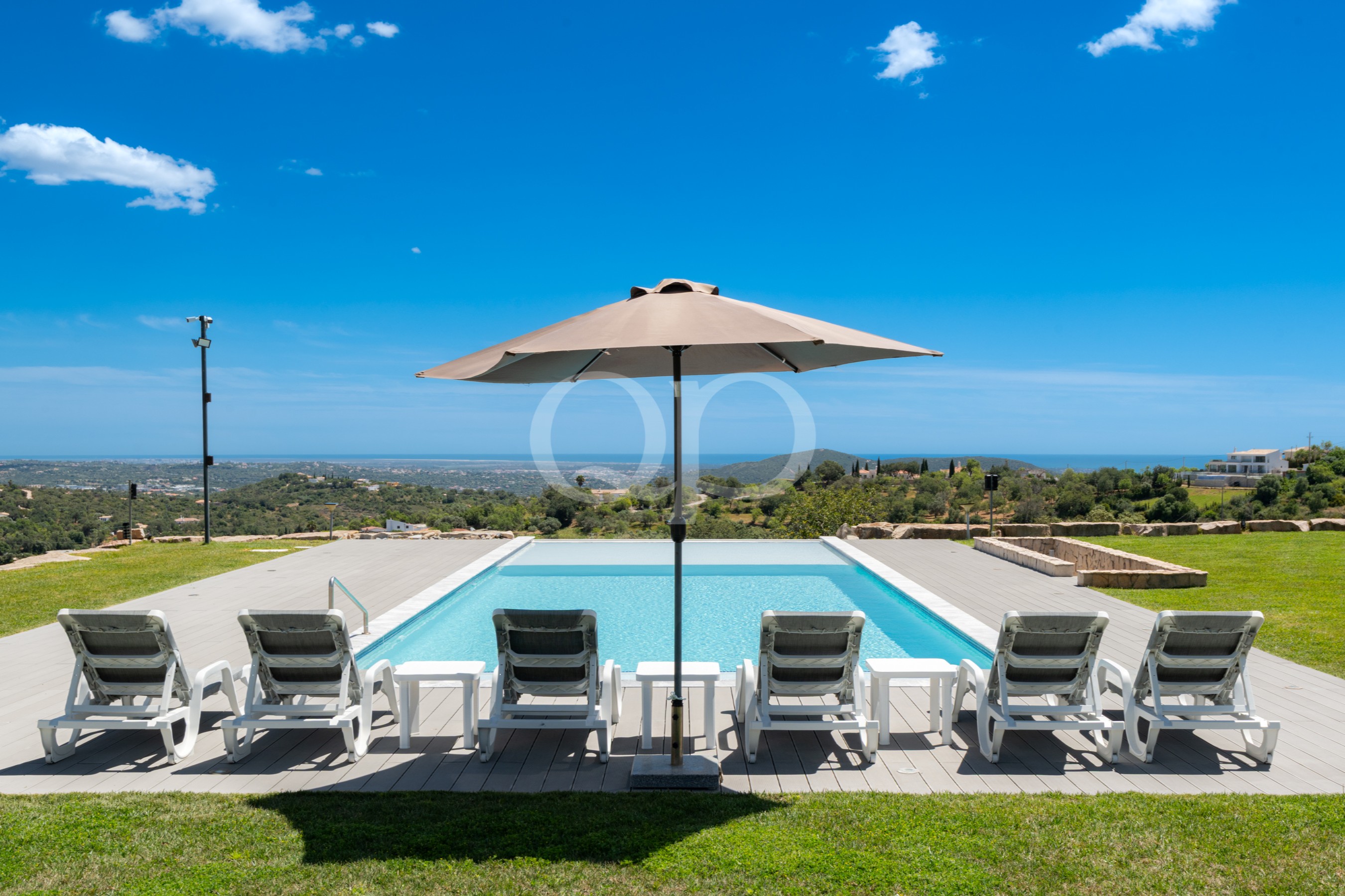 Poolside scene with white lounge chairs and a beige parasol, overlooking distant hills under a clear blue sky.