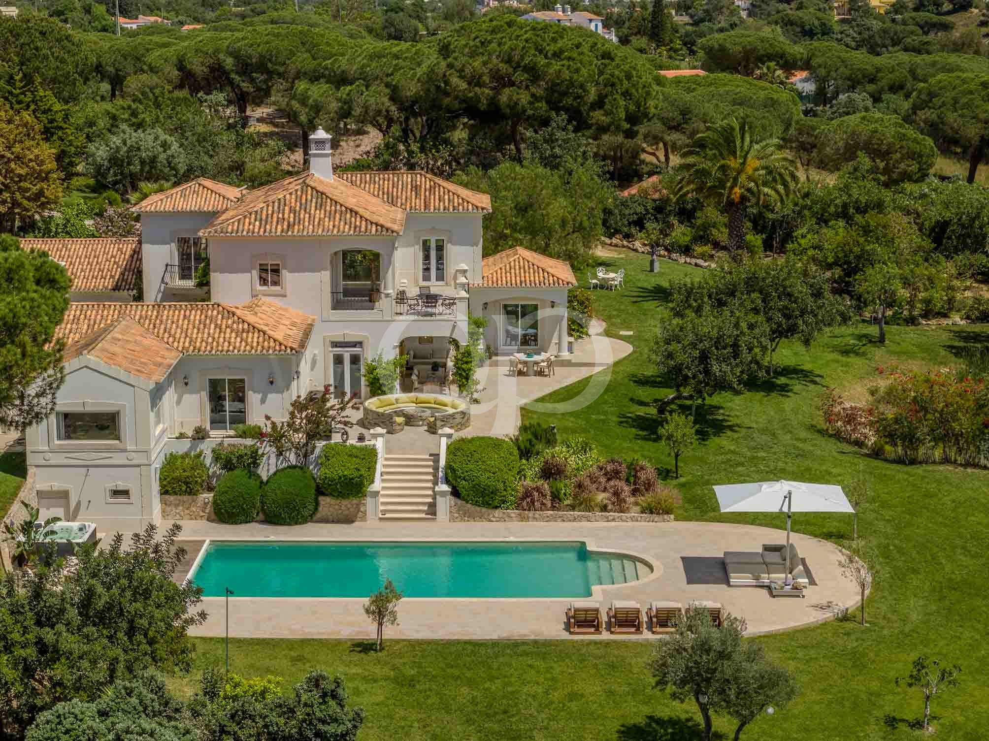 A white villa with a terracotta tile roof, surrounded by lush trees, a patio with seating, and a rectangular pool nearby.