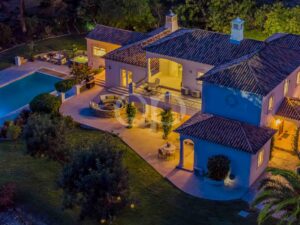 Aerial night view of a Mediterranean-style villa with blue-tiled roof, glowing windows, and an in-ground pool nearby.