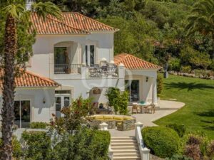 White Mediterranean villa with terracotta roofs, balcony seating, and surrounding palm trees in a lush garden setting.