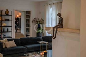Bronze sculpture of a seated figure perched on a railing in a modern living room, with a bookshelf and plants in the background.