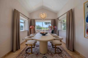 Bright dining room with a light-colored pedestal table surrounded by cream chairs, a glass bubble chandelier, and large windows with beige curtains.