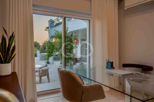 Living room with a glass dining table and brown chairs, white curtains, and a sliding glass door opening to a sunny patio with greenery outside.