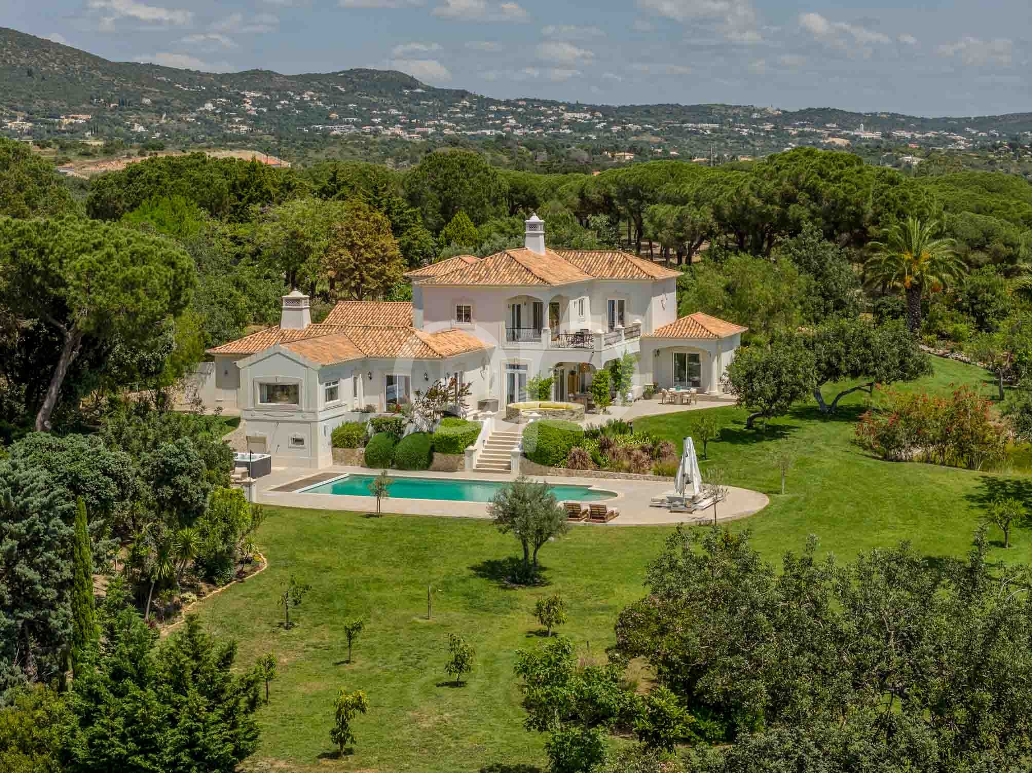 White Mediterranean-style villa with red-tiled roof, balcony, and a rectangular pool surrounded by a manicured lawn and trees, hillside backdrop.