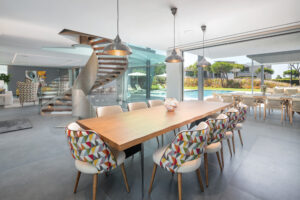 Offene Lobby mit einer spiralförmigen Metalltreppe, einem langen Holztisch und bunten Stühlen in geometrischen Formen; durch Glaswände hat man einen Blick auf den Außenpoolbereich.