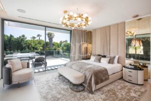 Ein luxuriöses Schlafzimmer mit einer großen Glaswand, die den Blick auf einen Balkon und üppiges Grün freigibt, einem beigen Polsterbett und einer dazu passenden Bank am Fußende.