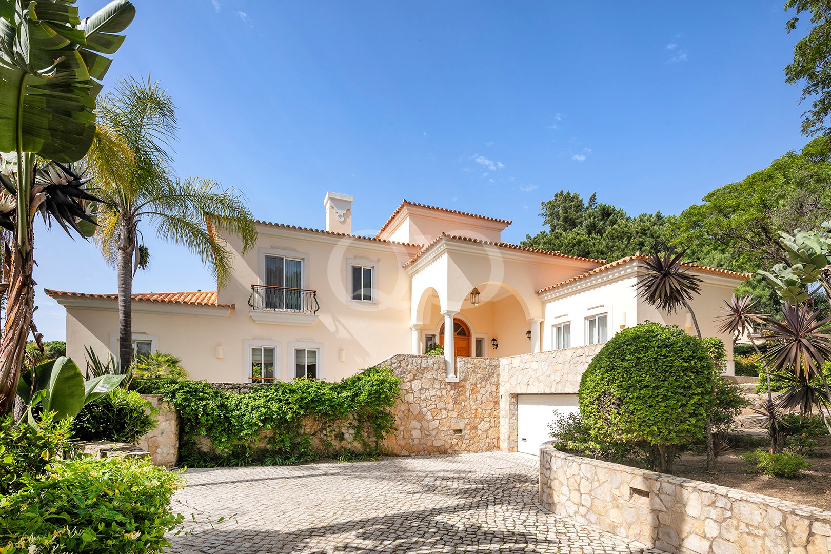 Mediterranean-style villa with peach stucco walls, terracotta roof, and arched entryway framed by a stone wall and lush landscaping under a blue sky.