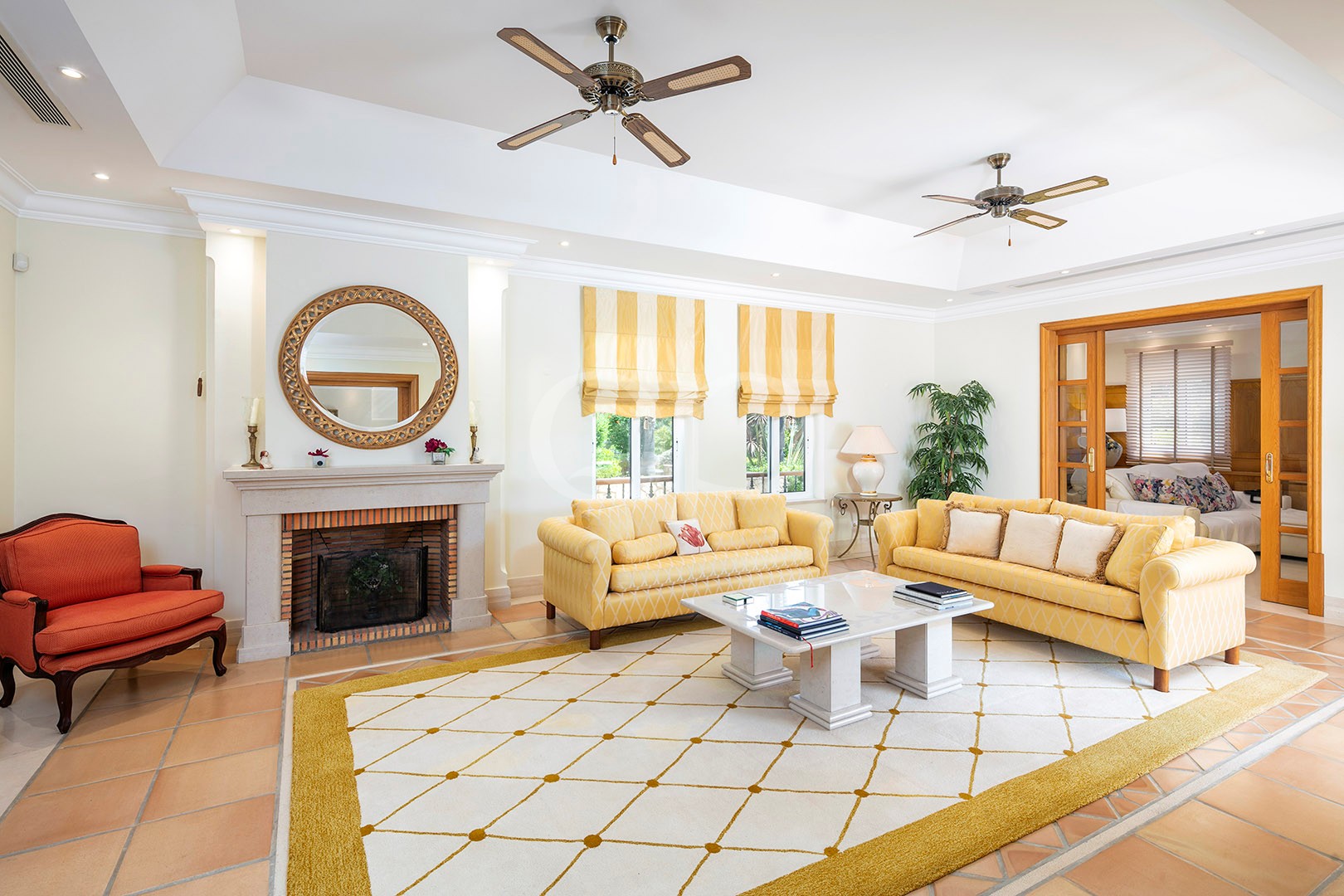 Bright living room with two yellow-patterned sofas, a white coffee table, and a brick-fireplace accented by a round wicker mirror over the mantel.