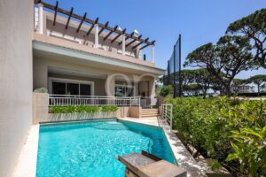 Modern beige house with a rectangular pool in the foreground, white railing, and a shaded patio area.