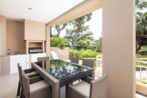 Outdoor patio with a glass-top dining table and wicker chairs, built-in grill, sink, and beige cabinetry under a shaded ceiling.